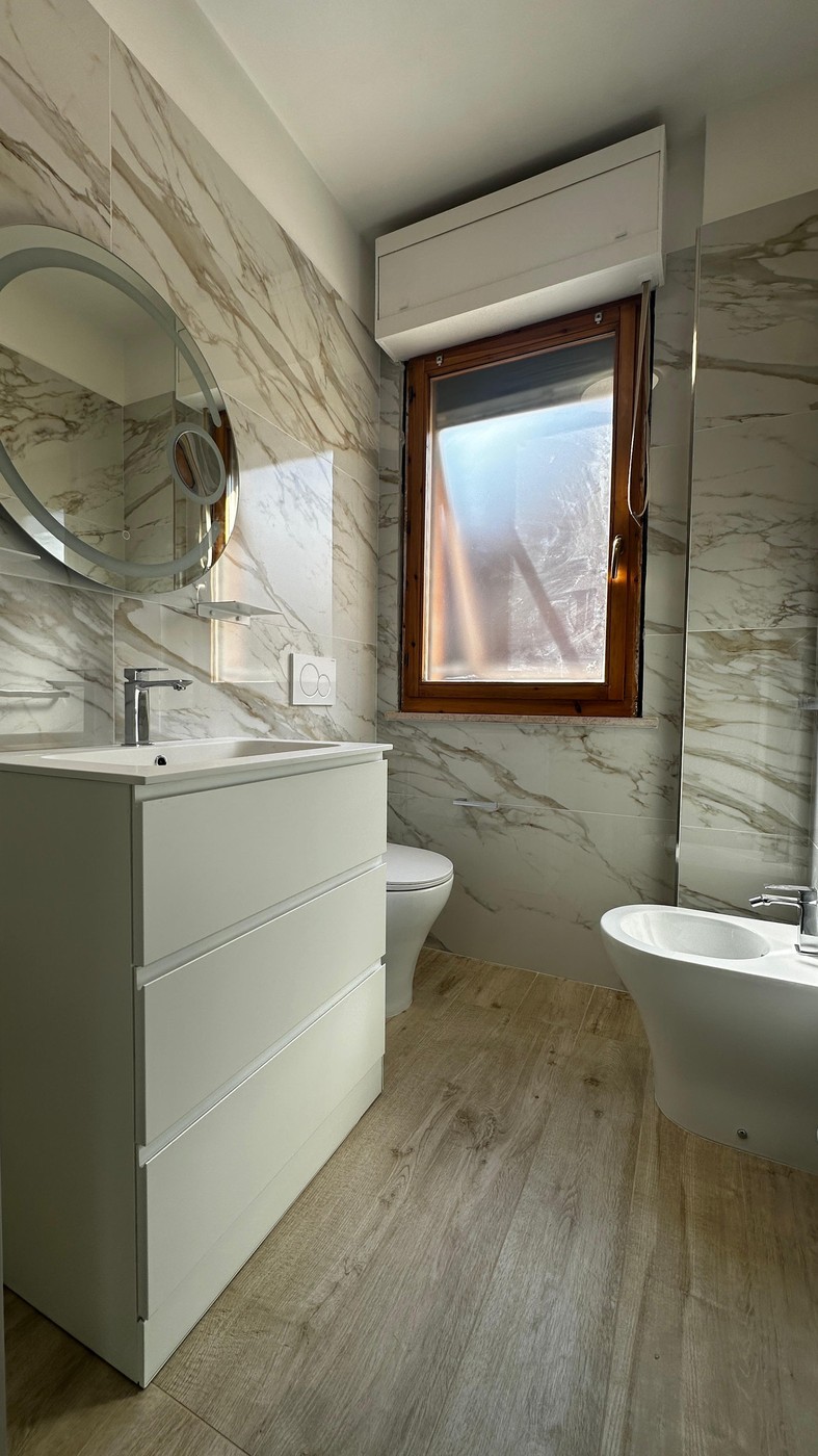 Vista frontale bagno calacatta: lavabo integrato, WC sospeso, bidet e specchio tondo — Senigallia 2026